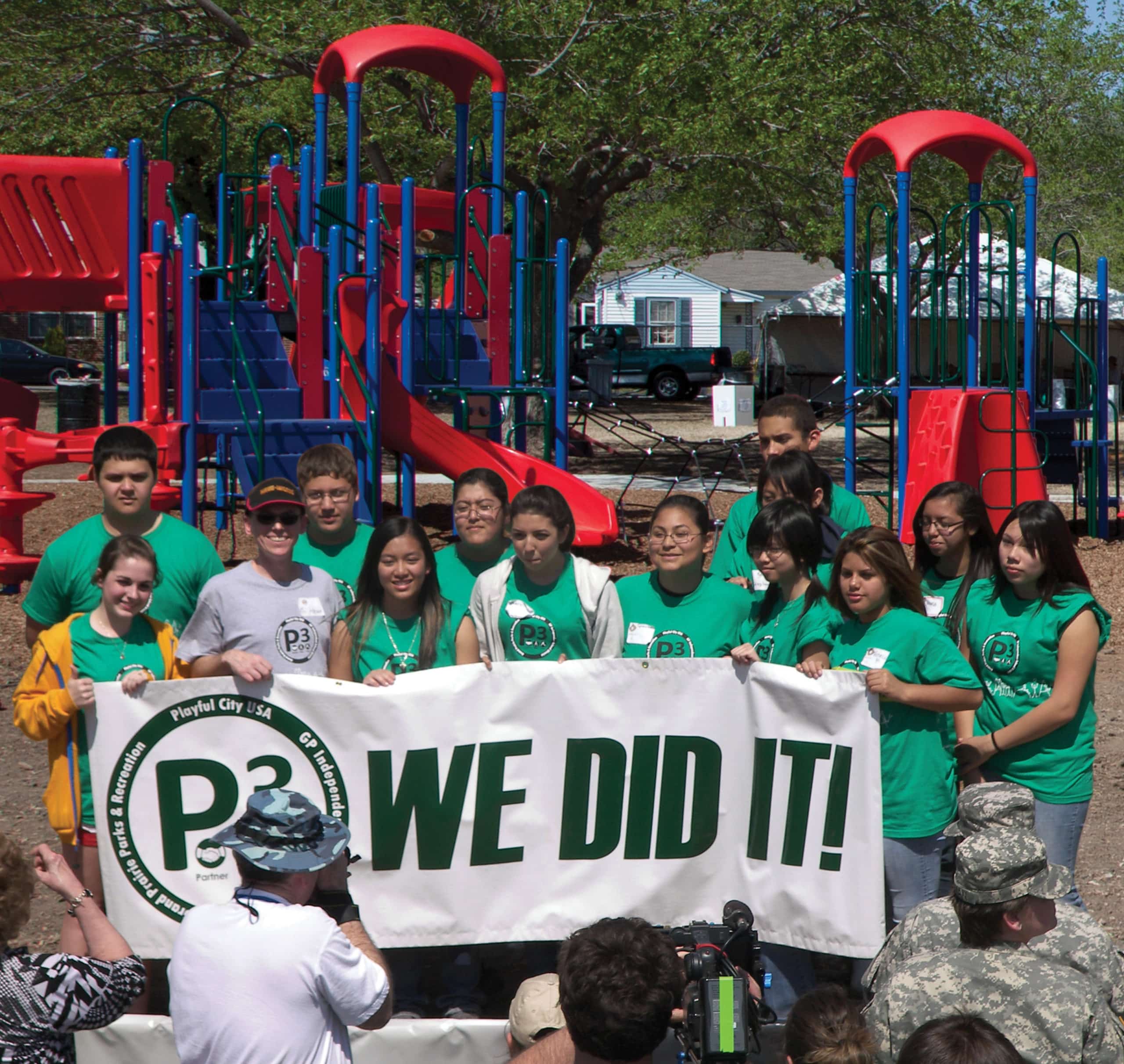 Several people wearing green shirts holding a banner that reads "P3 We Did It!" in front of an outdoor playground and a crowd
